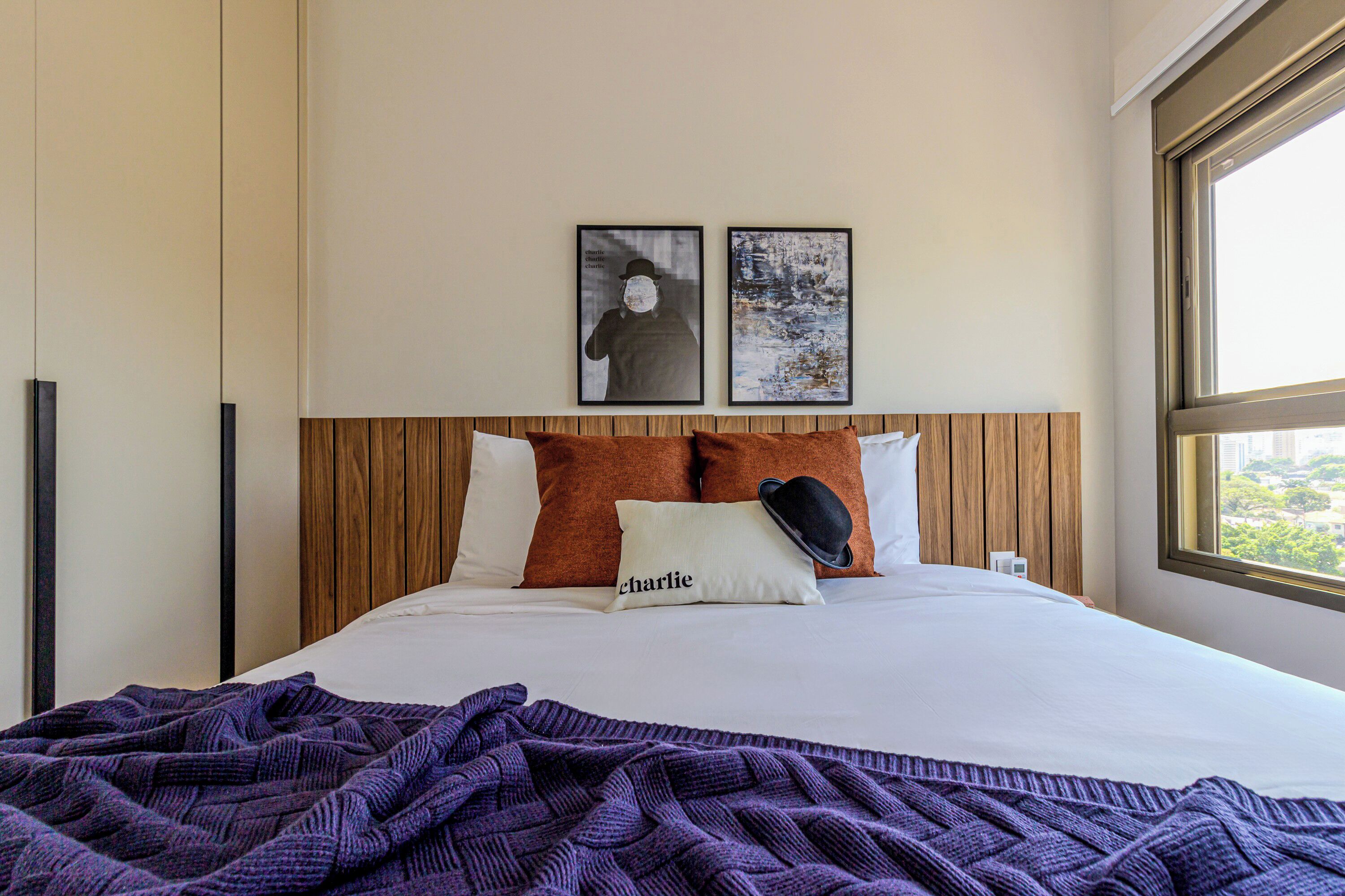 A neatly made bed with a purple blanket, two pillows with one labeled "Charlie," and two framed pictures on the wall.