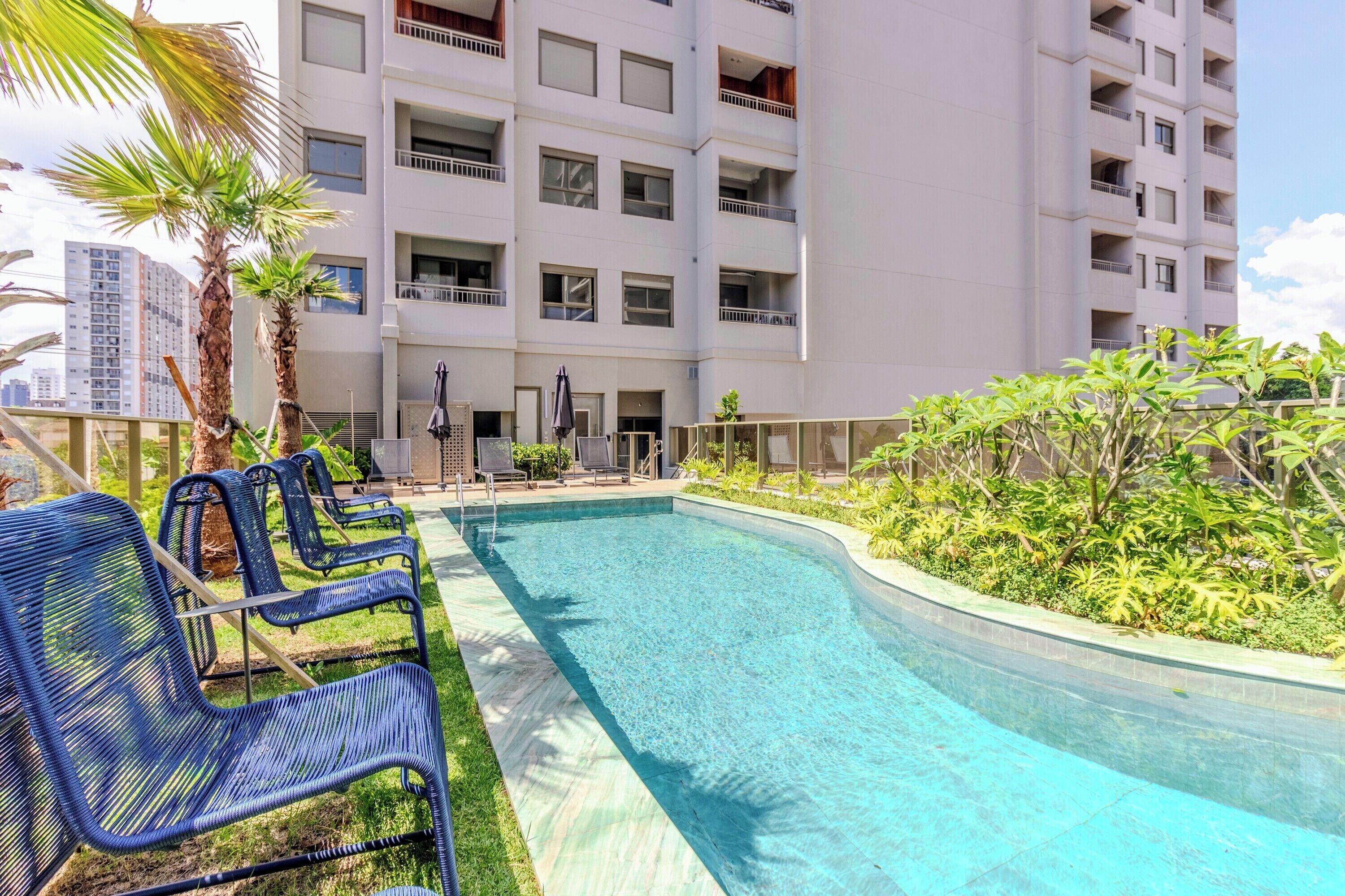A swimming pool with lounge chairs, surrounded by greenery and a multi-story building with balconies.