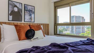 Ein Hotelzimmer mit großem Fenster, einem Bett mit weißen Leinentüchern, einer violetten Decke und zwei Kissen mit dem Namen "Charlie".