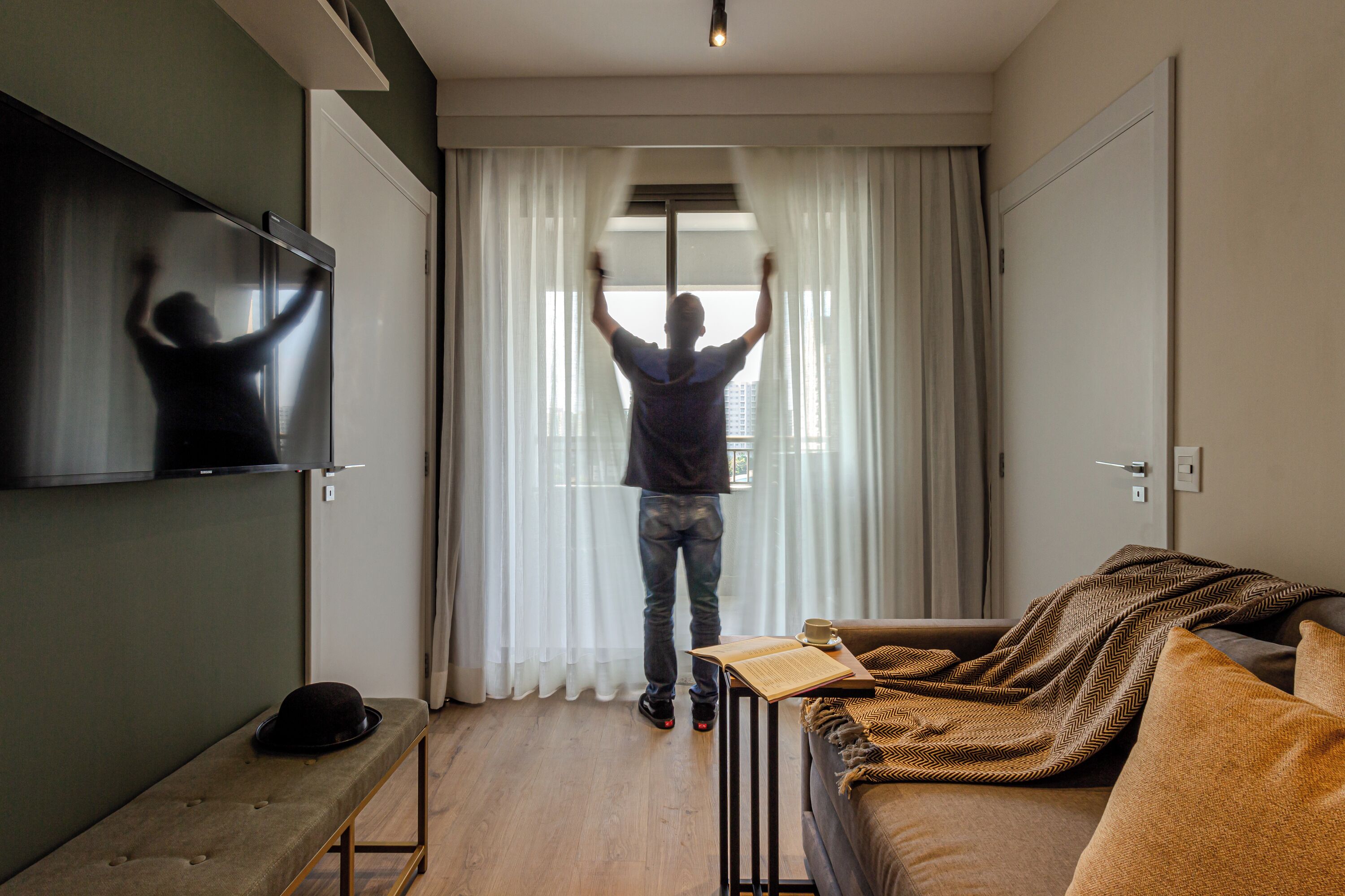 Ein modernes Wohnzimmer mit Sofa, Bank, Fernseher und einer Person, die am Fenster mit Vorhängen steht.
