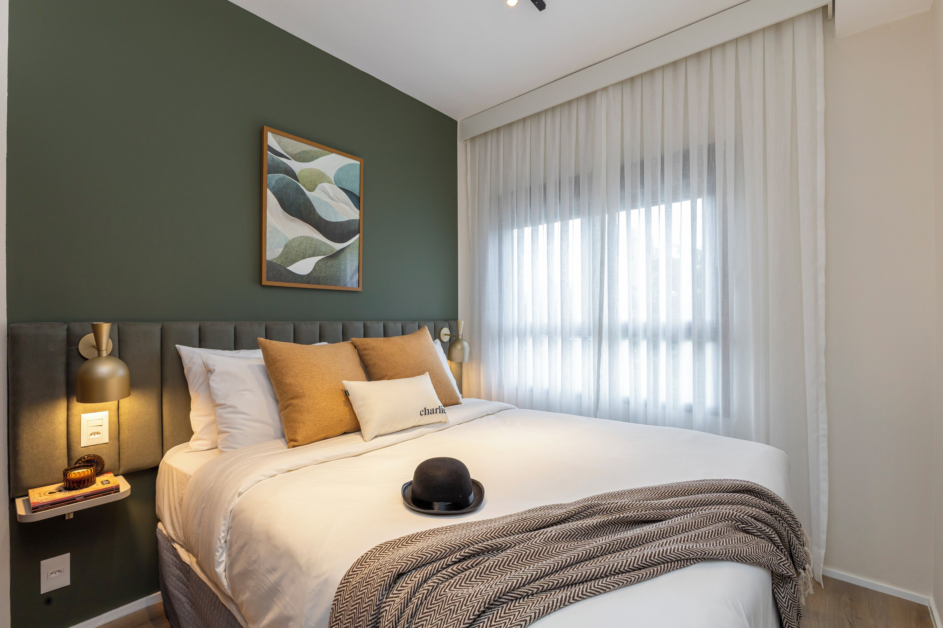 Uma cama bem arrumada com um manta estampada, um chapéu sobre a cama e uma luminária fixada na parede.