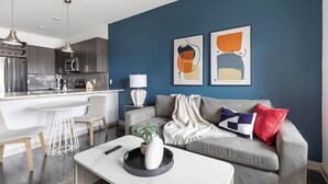 A modern living room with a grey sofa, a coffee table, and a kitchen area with a bar and dining space. - Landing | Stylish 1BD, Gym, Pool (McKinney)