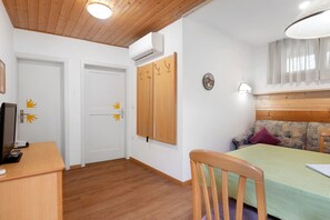 A room with a wooden ceiling, a bed with a green bedspread, a wooden desk with a computer, a couch, and a wall-mounted air conditioner.