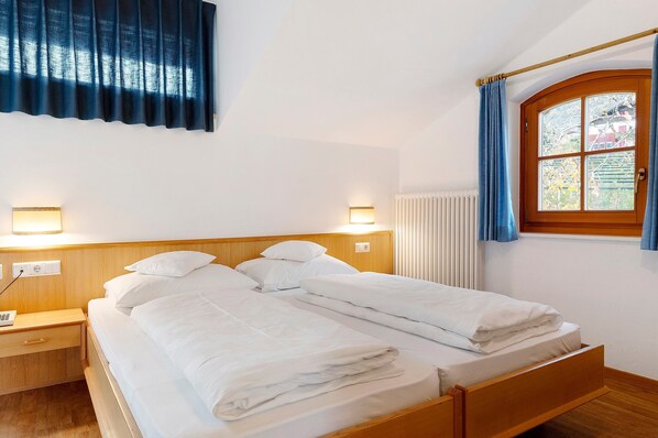 Un lit double avec du linge de lit blanc, une tête de lit en bois, des lampes de chevet, une fenêtre avec des rideaux et un radiateur.