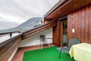 Un balcon agrémenté d’une moquette artificielle verte, d’une chaise en métal, d’une table recouverte d’une nappe jaune et offrant une vue sur une montagne.