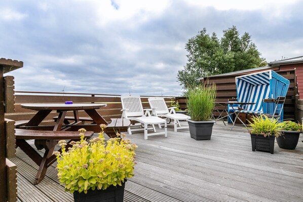 Una terraza de madera con una mesa de picnic, sillas de lounge blancas y plantas en macetas.