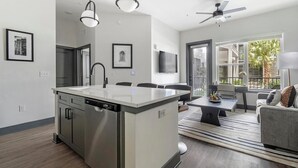 A modern kitchen with a central island, a dining area with a sofa and table, and a balcony with a view of palm trees.
