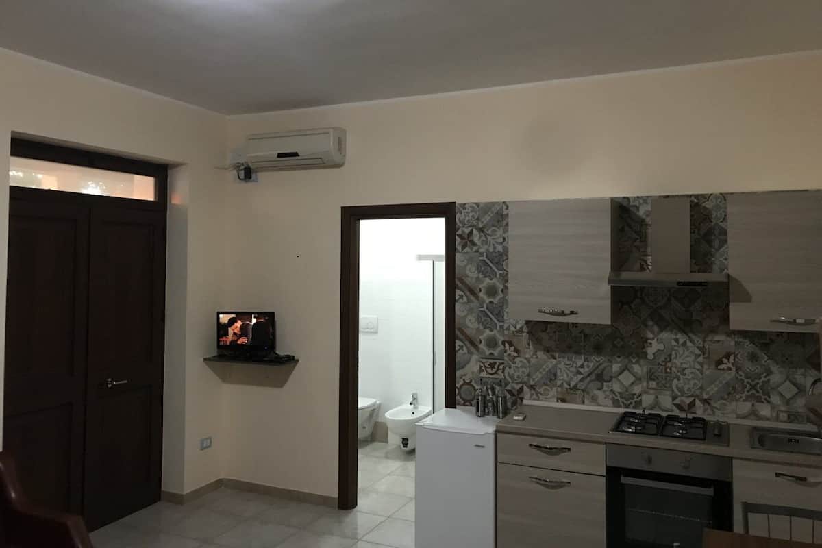 Una cocina con estufa, horno y salpicadero de azulejos. Hay un televisor en un estante y una puerta que da a otra habitación.