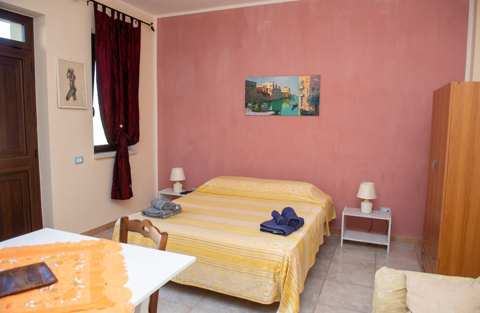 A bedroom with a bed, two bedside tables, a wooden wardrobe, a window with curtains, and a painting on the wall.
