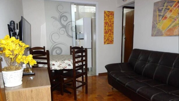 A living room with a wooden dining table, two chairs, a black leather sofa, and a potted plant with yellow flowers.