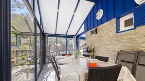 A covered outdoor dining area with a table set for four, wicker chairs, and a view of the garden through large windows. - Holiday Home in Carcans – Terrace, Air Conditioning, Near Lake (Carcans Maubuisson)