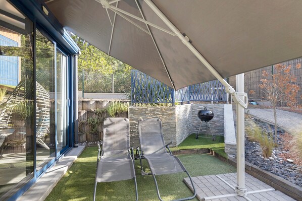 A covered outdoor seating area with two lounge chairs, a stone wall, and a grill.
