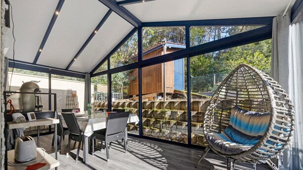 A modern outdoor dining area with a glass wall, a wicker chair, and a table with chairs. - Holiday Home in Carcans – Terrace, Air Conditioning, Near Lake (Carcans Maubuisson)