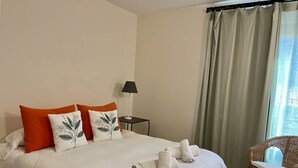 A neatly made bed with white and orange pillows, a small table with a lamp, and a wicker chair. - Villa Over the 5th Green (San Bartolomé de Tirajana)