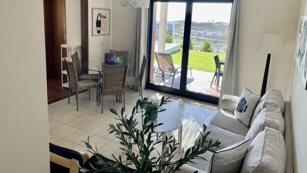 A living room with a sofa, a dining table with chairs, a small table with a plant, and a view of a pool and greenery outside. - Villa Over the 5th Green (San Bartolomé de Tirajana)