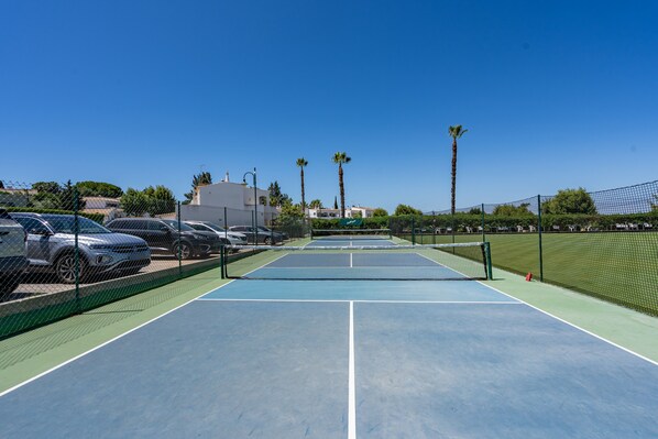 An outdoor tennis court with a net, surrounded by a fence, and adjacent to parked cars.
