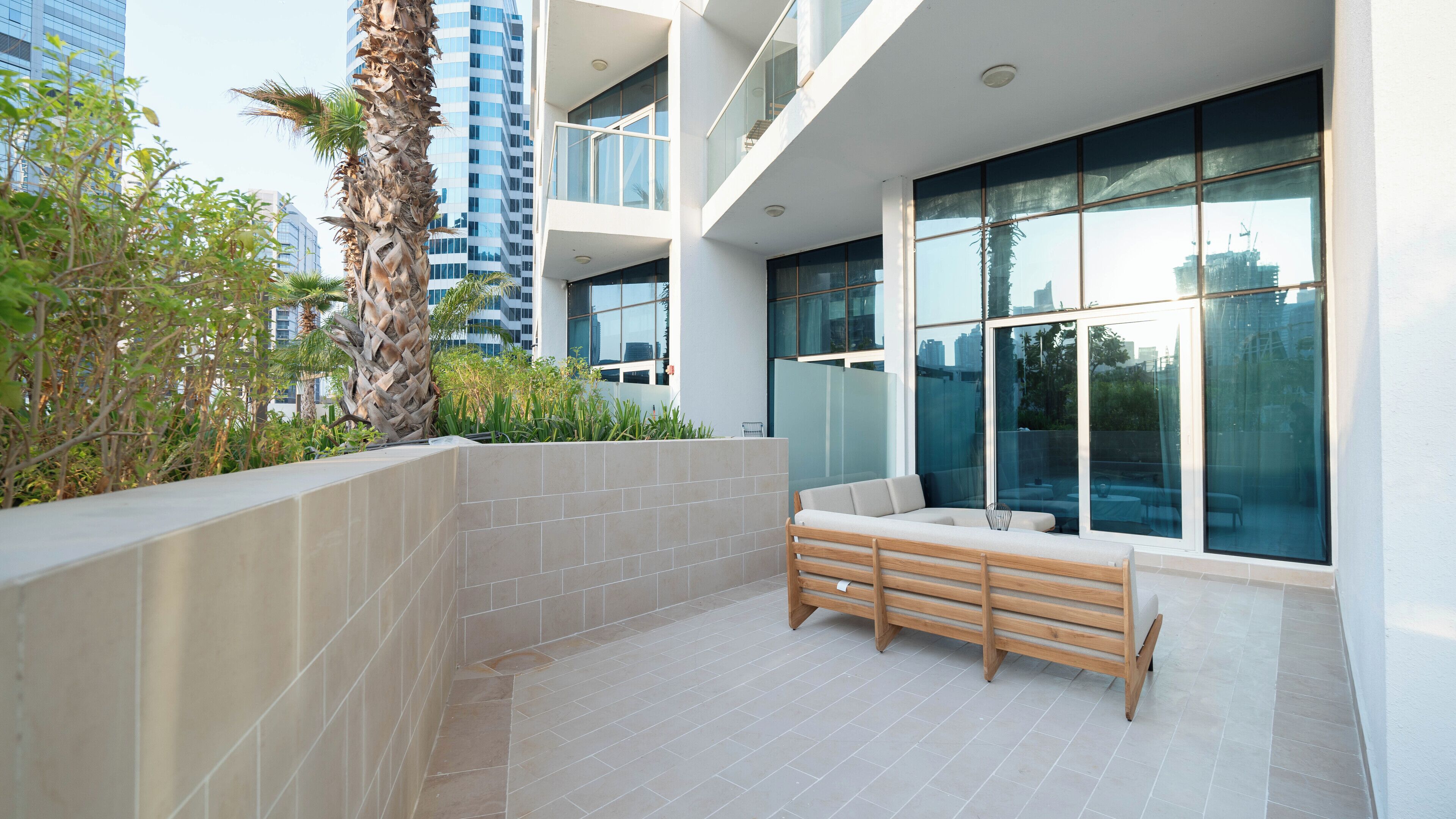 A modern outdoor seating area with a bench, surrounded by a low wall and plants, with a large window offering a view of the cityscape.