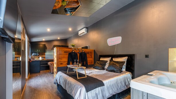 A modern bedroom with a large bed, a wooden wardrobe, a small table, a hot tub, and a view into the kitchen.