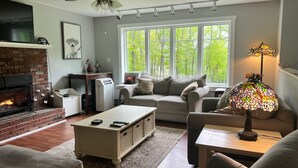 A living room with a fireplace, a ceiling fan, a large window with a view of trees, and a table lamp.