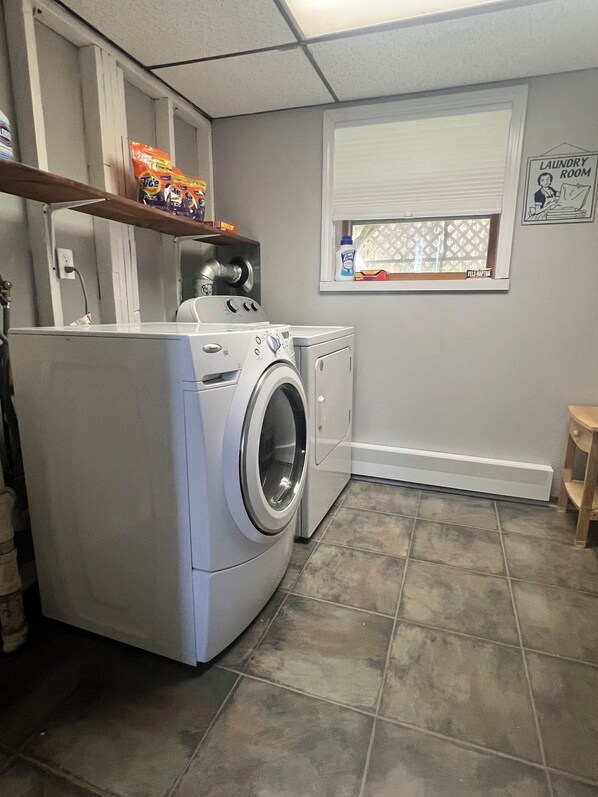 Une buanderie équipée d’un lave-linge et d’un sèche-linge à chargement frontal, d’une fenêtre avec des stores et d’une étagère contenant des produits de lessive.
