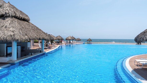 A swimming pool with lounge chairs and thatched umbrellas, surrounded by a beach and ocean view.