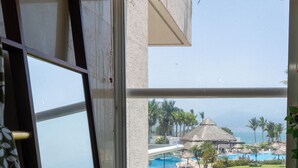 A room with a large window overlooking a pool area, a beach, and a thatched-roof structure.