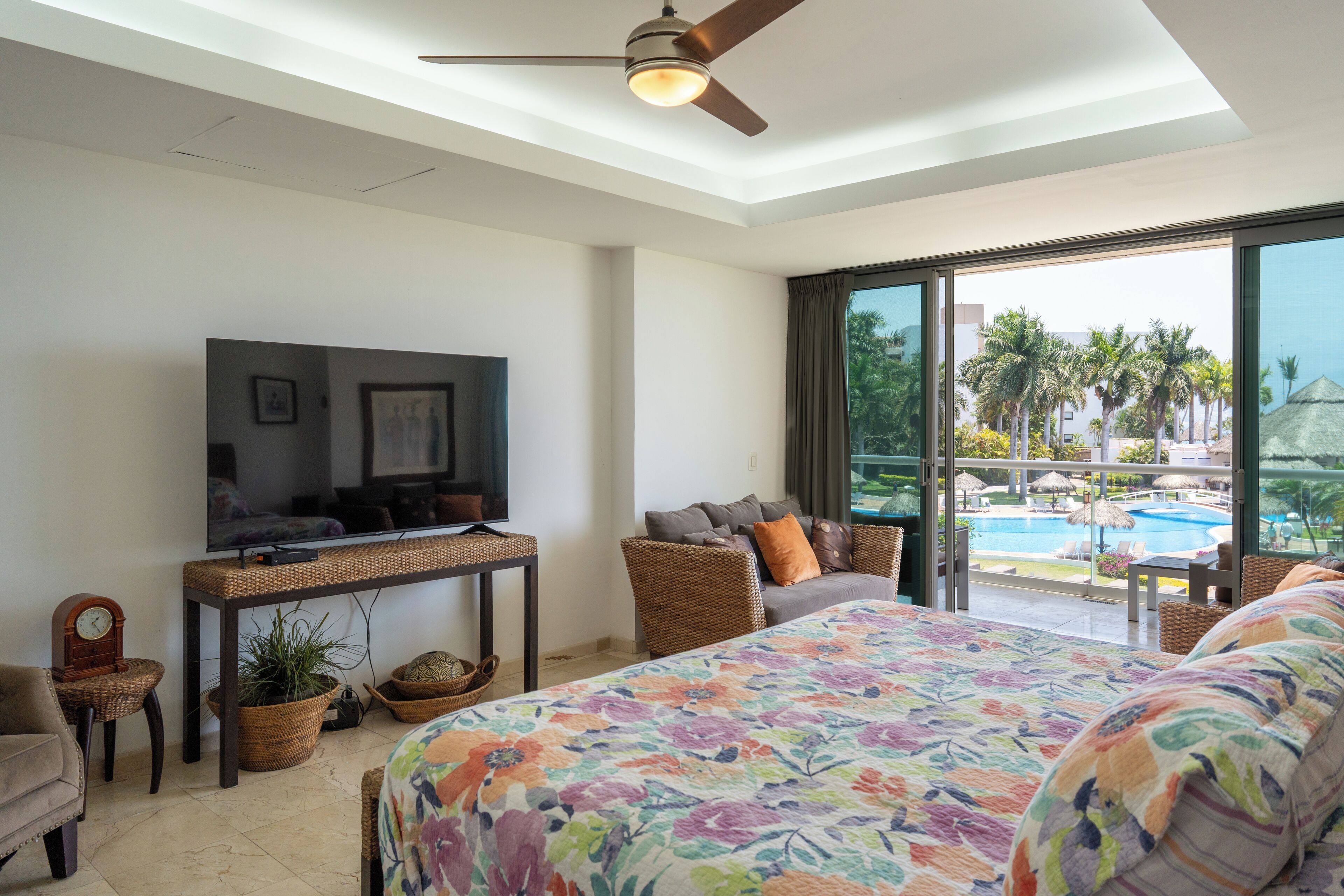 A bedroom with a bed, a television, a ceiling fan, a window with a view of a pool and palm trees.