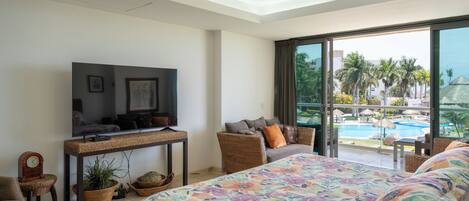 A bedroom with a bed, a television, a ceiling fan, a window with a view of a pool and palm trees.