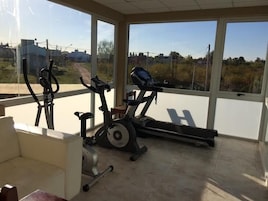 Uma academia em casa com duas bicicletas ergométricas e uma esteira, grandes janelas com vista para uma paisagem rural.