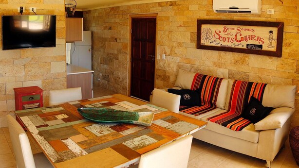 A cozy living room with a wooden table, a sofa with colorful blankets, a flat-screen TV mounted on the wall, and a framed sign above the sofa. - Casa Río Spa (San José)