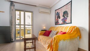 A room with a sofa, a table, a window with a view of buildings, an air conditioning unit, and a wall with a framed picture. - Brigth Apartment 'Macarena' with Balcony, Wi-Fi and Air Conditioning (Sevilla)
