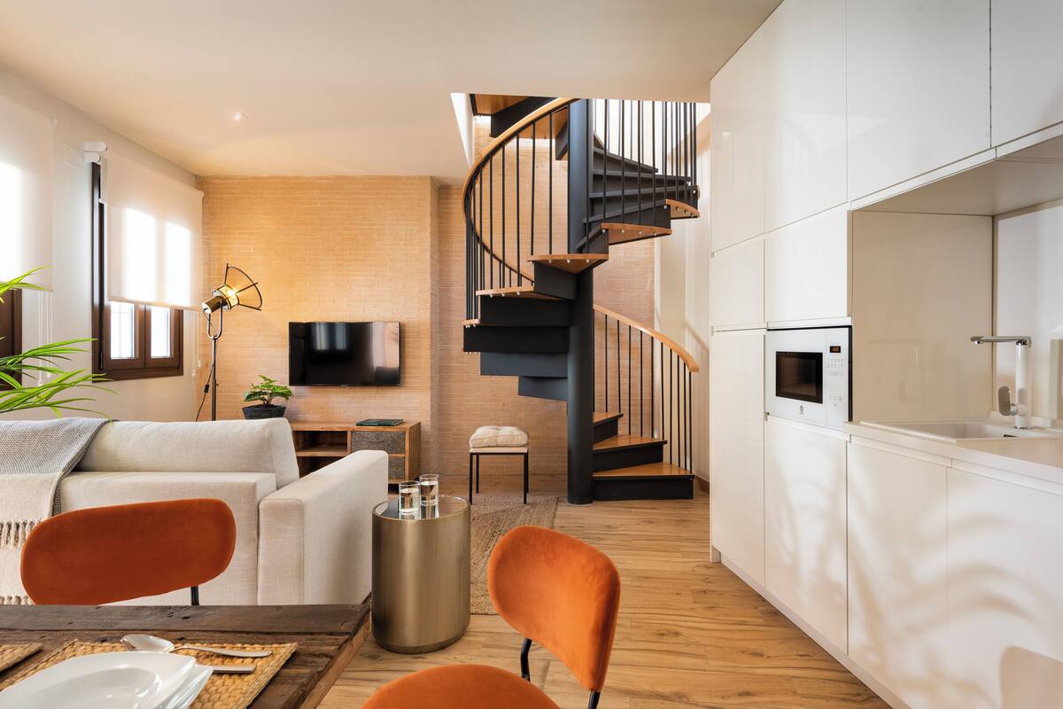 A modern living room with a sofa, two orange chairs, a dining table with plates, a TV, and a spiral staircase leading to a kitchen area.