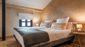 A bedroom with a brick wall, a large bed with multiple pillows, a bedside table with a lamp, and a window with blinds. - MH Apartments Central Sevilla (Seville)