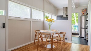 Ein Essbereich mit einem runden Tisch, sechs Stühlen, einer Vase mit gelben Blumen und Blick ins Freie durch große Fenster.