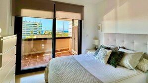 A bedroom with a bed, a nightstand, a ceiling fan, and a view of buildings and a body of water through a sliding door.