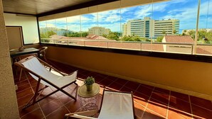 A balcony with two lounge chairs, a small round table with a plant, and a view of a cityscape with buildings and a clear sky.