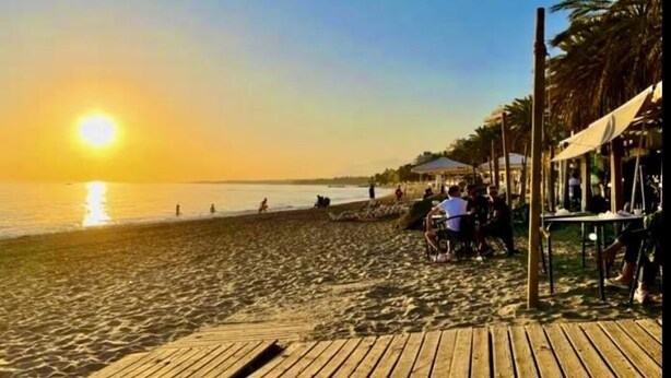 A beach with a wooden walkway leading to a covered area with tables and umbrellas.