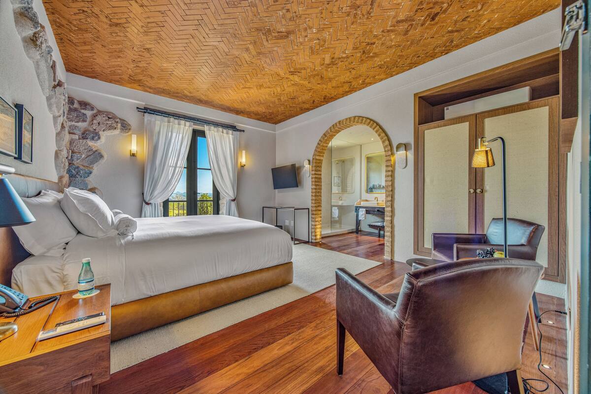 A hotel room with a wooden ceiling, a large bed, a sitting area with a chair, and a bathroom visible through an archway.