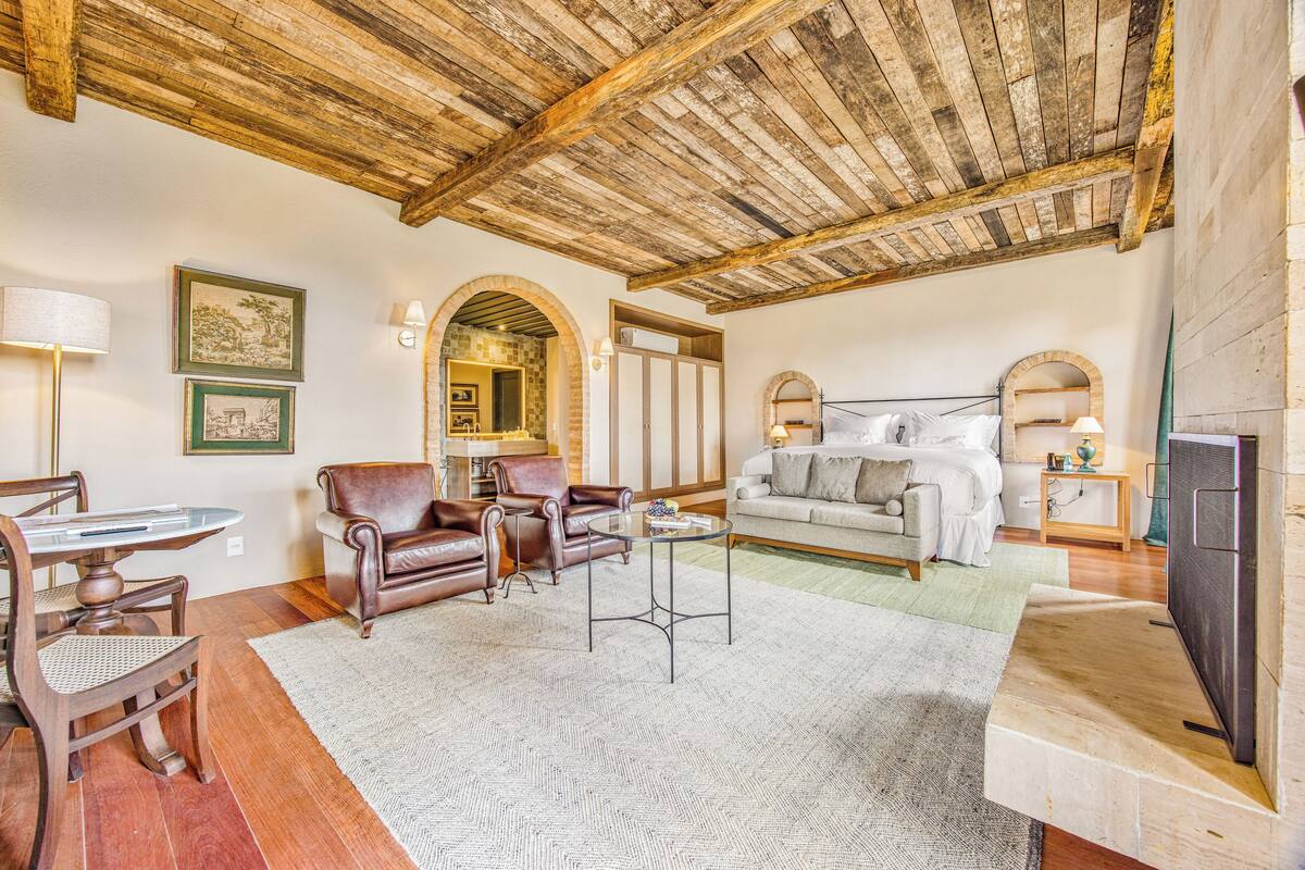 A spacious living room with a wooden ceiling, a fireplace, a dining area with a table and chairs, and a cozy seating area with a sofa and armchairs.