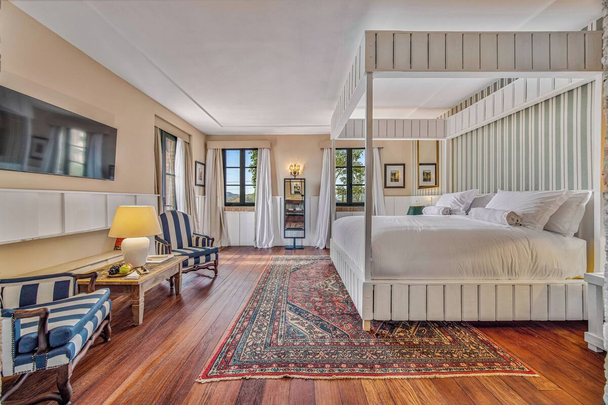 A bedroom with a four-poster bed, a wooden floor, a large rug, a television, and a desk with a lamp.