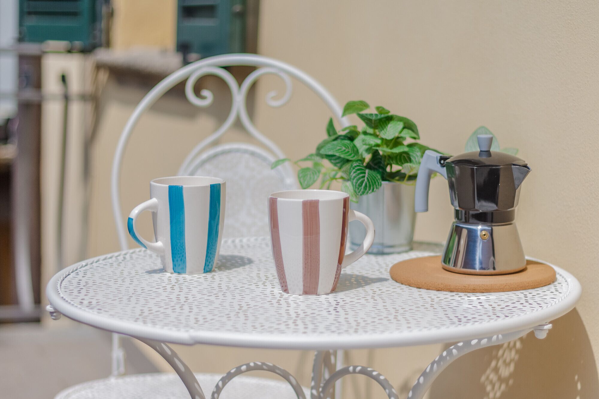 Una mesita redonda con dos tazas a rayas y una cafetera sobre una silla de metal blanca.