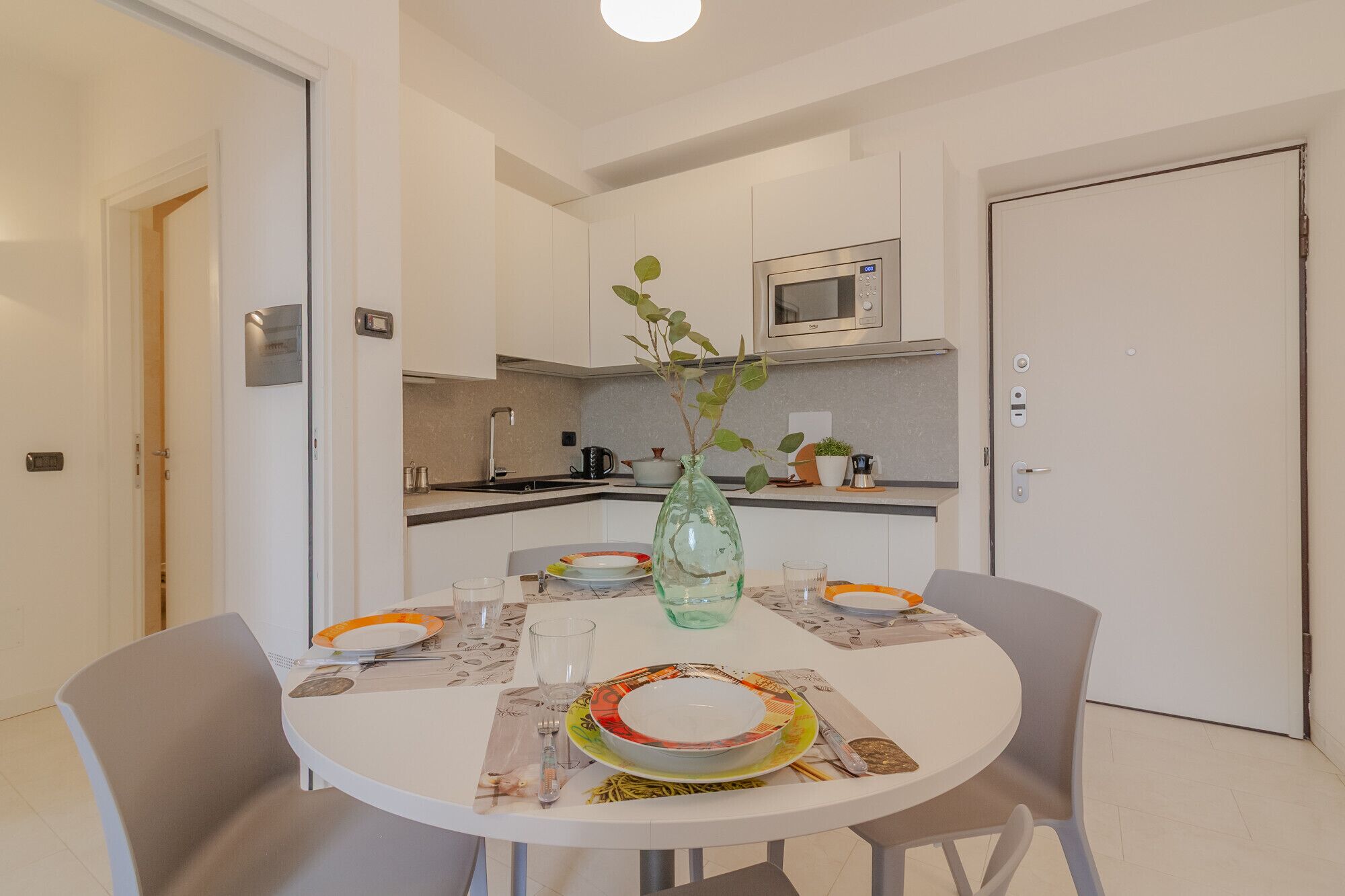 A compact kitchen with white cabinets, a microwave, and a dining table set for two with plates and glasses.