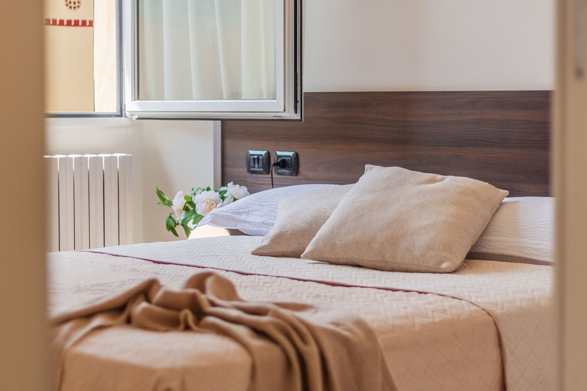 Ein ordentlich gemachtes Bett mit einer beigen Decke und Kissen, ein Nachttisch mit Blumen in einer Vase und ein Fenster mit Vorhängen.