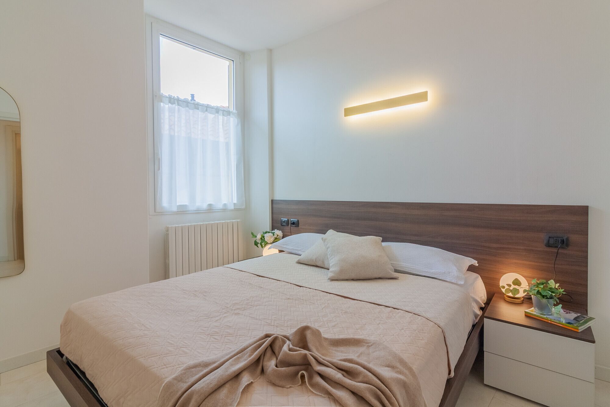Ein ordentlich eingerichtetes Schlafzimmer mit einem Bett, Nachttischen, einer Wandleuchte und einem Fenster mit durchsichtigen Vorhängen.