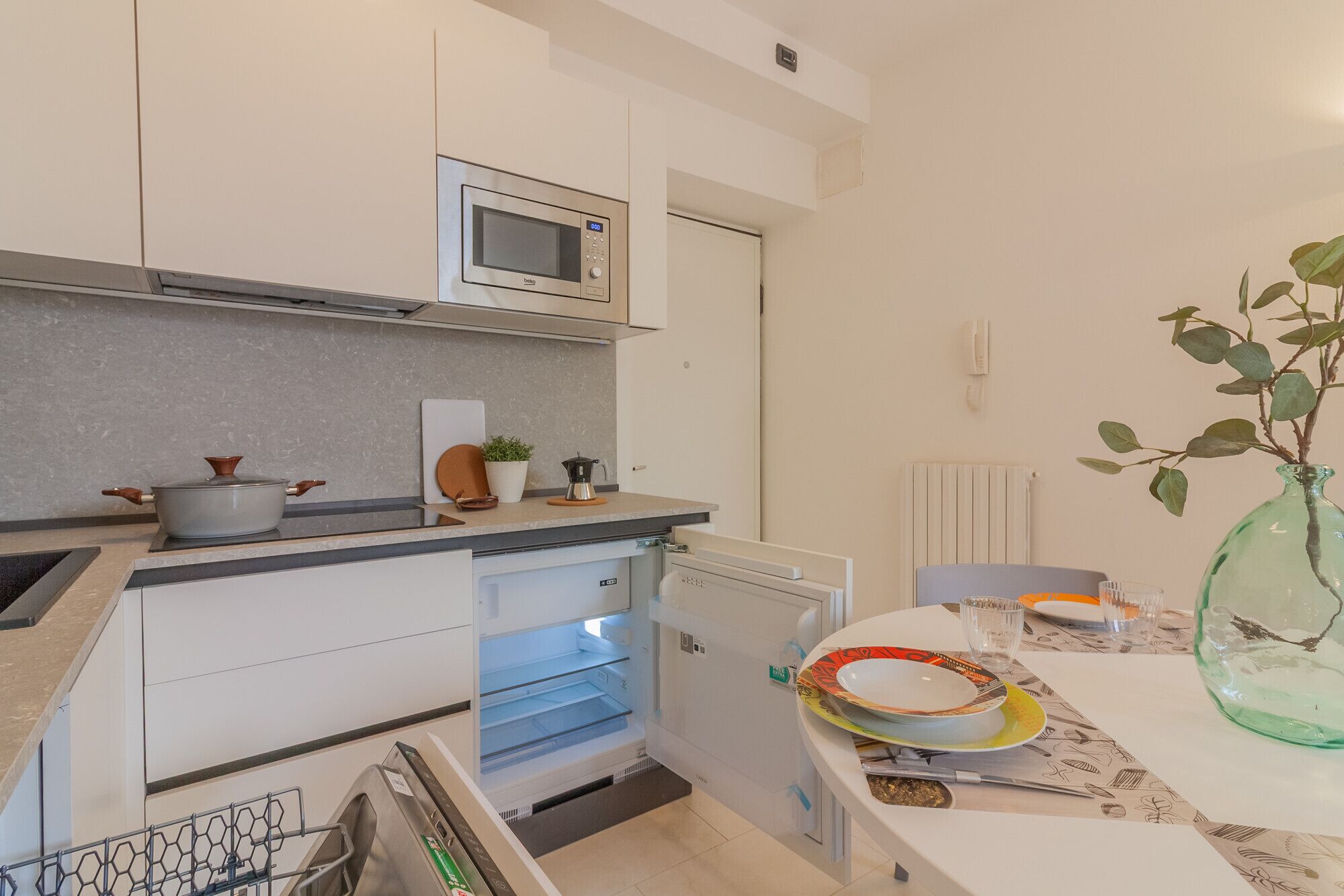 A compact kitchen with a microwave, stove, and refrigerator. There's a small dining area with a table set for two and a vase with green leaves.