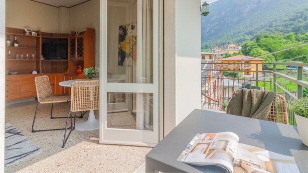 Un balcón con vistas a un paisaje montañoso, una zona de comedor con mesa y sillas, y una estantería con diversos objetos.