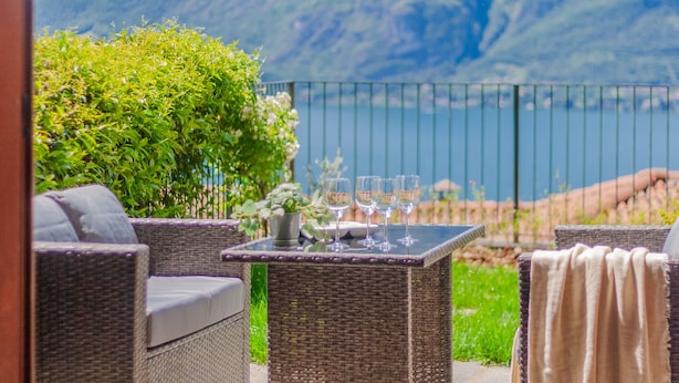 Eine Terrasse mit Korbmöbeln, einem Tisch mit Weingläsern und Blick auf einen Berg und einen See.