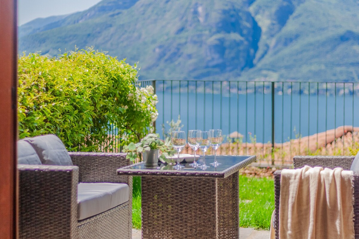 A patio with wicker furniture, a table with wine glasses, and a view of a mountain and lake.