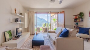 Ein modernes Wohnzimmer mit Sofa, Couchtisch und Fernseher. Es gibt einen Balkon mit Blick auf Berge und einen Garten.