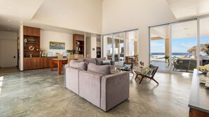 A spacious living area with a large grey sofa, a dining table with chairs, and a view of the beach through large glass doors. - Kas di Bon Beis - Spacious Oceanfront Villa with Full A/C (Kralendijk)